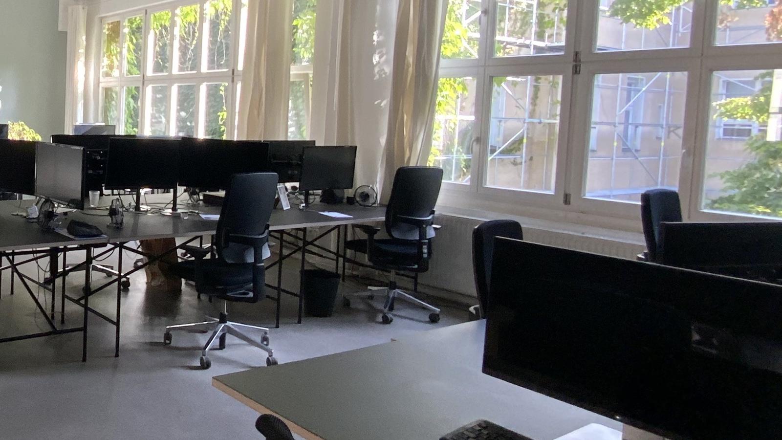 Empty office desks with computer monitors and black office chairs beside tall windows on a sunny day
